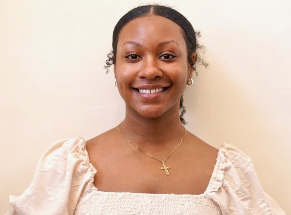A young woman smiles while wearing a gold cross necklace, photographed in a bright, neutral setting.
