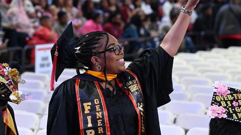 A graduate in academic regalia raises her arm in celebration during a commencement ceremony, surrounded by seated attendees.