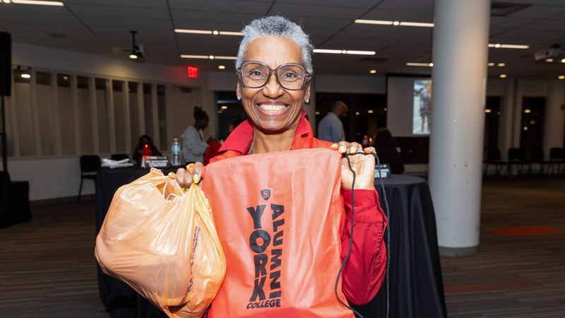 A smiling older woman wearing glasses holds a large orange bag with 'YORK COLLEGE' printed on it, standing indoors at an institutional advancement gathering.