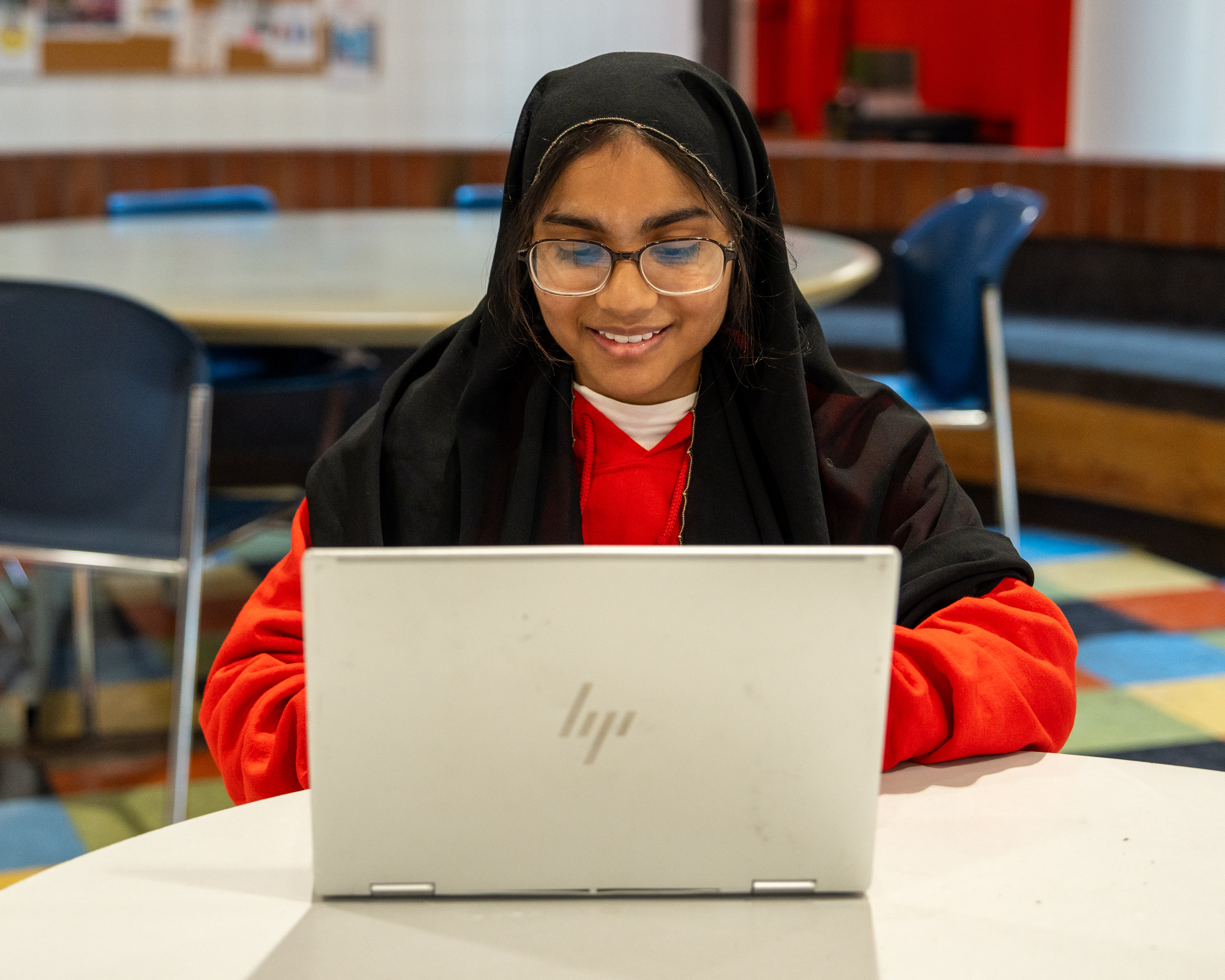 Student wearing glasses and a headscarf uses a laptop while studying at a table in a campus learning space.