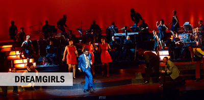 Picture of man with blue suit dancing in front of three Dreamgirls dressed in red all in front of large band with red backdrop.
