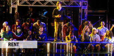 Singer standing on top of long metal table surrounded by others lit in blue light.